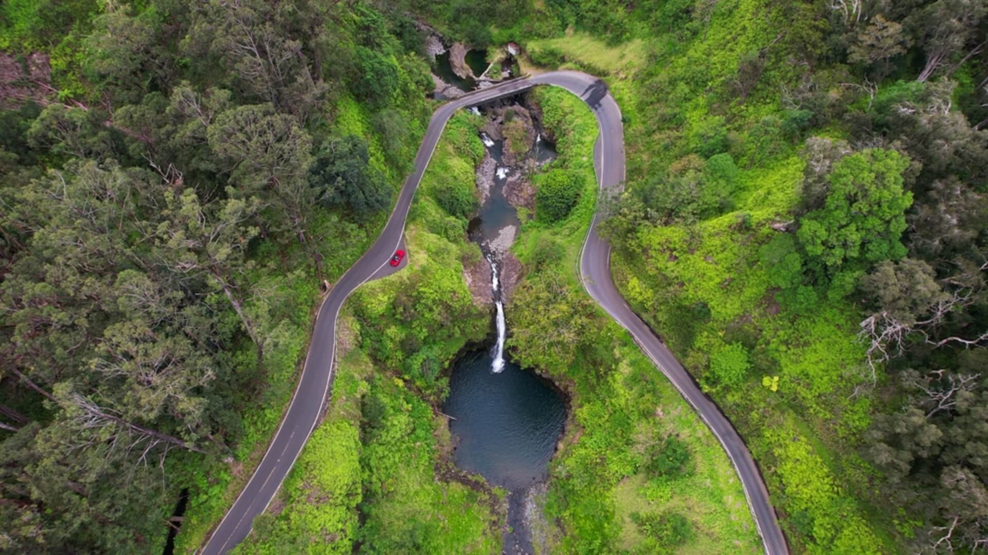 An overhead shot of a car driving around a bend on the Road to Hana near a waterfall, one of the most scenic drives on Maui